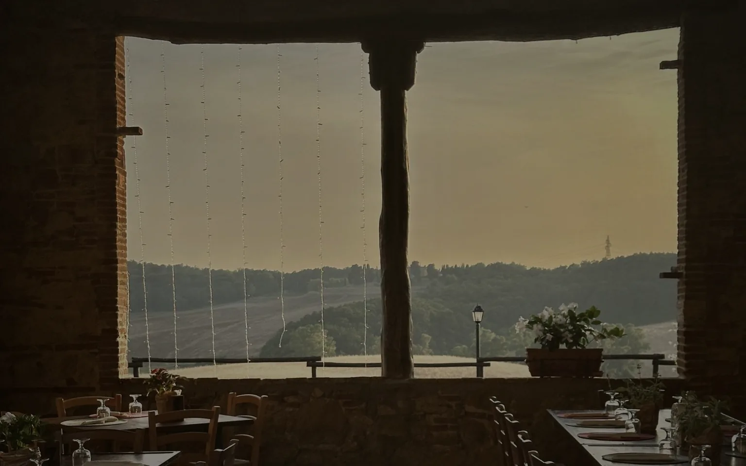 A dining room at dusk seen through a wide stone arch, tables laid, hills and water beyond.
