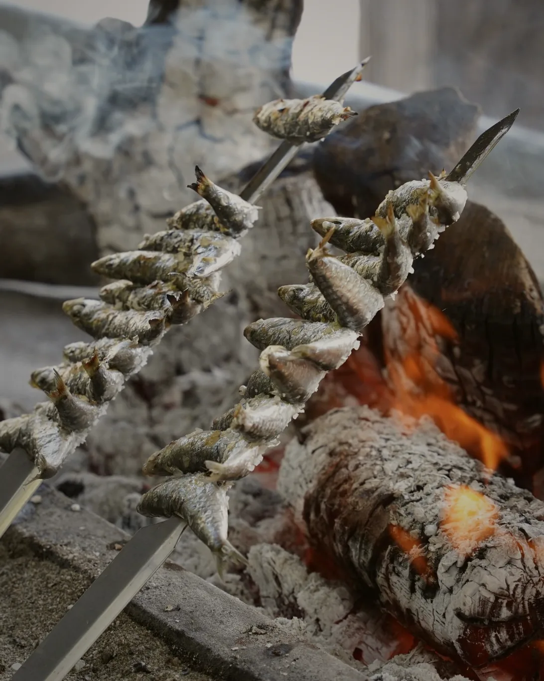 Sardines threaded onto cane skewers, propped at an angle beside an open driftwood fire and cooked in the smoke.